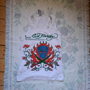 Vit Ed Hardy linnetopp med dödskalletryck - Säljer en vit linnetopp från Ed Hardy med stort färgglatt tryck framtill som visar en blå dödskalle omgiven av eldflammor och rosor. Toppen har brottarrygg och grönt logotryck både fram och bak.