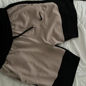 Nike shorts i beige och svart - Snygga shorts från Nike i beige och svart med svart snörning i midjan och klassisk Nike-logga på vänster sida. Perfekta för träning eller avslappnade dagar. Mjukt material och elastisk midja för extra komfort.Storlek M