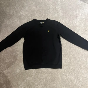 Svart sweatshirt från Lyle & Scott Junior - Säljer en svart sweatshirt från Lyle & Scott Junior med klassisk rund halsringning och den ikoniska gula loggan broderad på bröstet. Perfekt basic tröja med lång ärm och mjukt material.