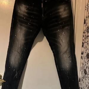 Svarta jeans med slitningar från Dsquared2 - Säljer ett par svarta jeans från Dsquared2 med coola slitningar och vita färgstänk. Jeansen har en smal passform och klassisk femficksdesign. Märkeslapp bak i midjan.priset är inte hugget i sten och byxrona är som nya