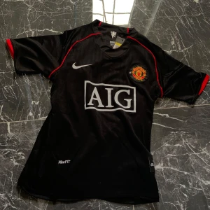 Manchester United Ronaldo 7 svart fotbollströja - Säljer en svart Manchester United fotbollströja med röda detaljer, AIG sponsortryck och klubbmärke på bröstet. På ryggen står det 'Ronaldo' och nummer 7. Tröjan är kortärmad och har NikeFIT-material för bra ventilation.
