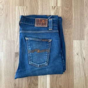 Blå jeans från Nudie Jeans - Säljer ett par klassiska blå jeans från Nudie Jeans med orange sömmar och patch bak. Modellen har raka ben och normal passform. Jeansen har lagade partier på benen och tydlig slitning för en cool, använd look.