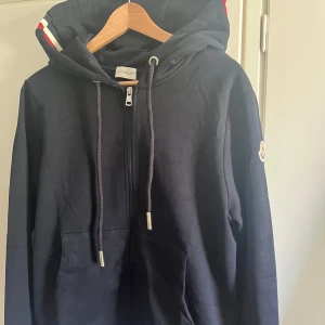 Mörkblå kofta från Moncler - Säljer en mörkblå kofta  från Moncler med dragkedja och huva. Jackan har Moncler-logga på ärmen och snygga ränder i rött och vitt på huvan. Tillverkad i mjuk bomull och har fickor framtill. 10/10 skick och skickas i moncler påse. QR och NFC funkar!