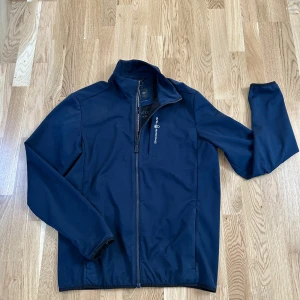 Marinblå softshelljacka från Sail Racing - Säljer en marinblå softshelljacka från Sail Racing med dragkedja framtill och hög krage. Jackan har broderad logga på bröstet och svart panel över ryggen. Perfekt för blåsiga dagar och enkel att matcha med det mesta.