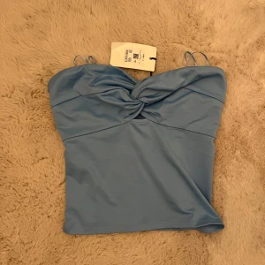 Ljusblå croppad tubtopp med twist framtill - Säljer en ljusblå croppad tubtopp från Bik Bok med twistad detalj framtill och smala axelband. Baksidan har smock för extra komfort och passform. Perfekt för sommardagar och enkel att matcha med jeans eller kjol.