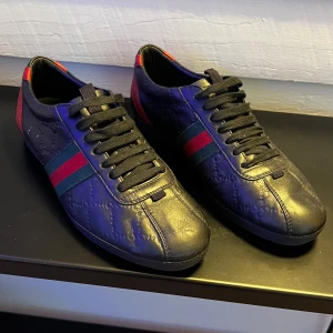Gucci Skor LÄS BESKRIVNING - Säljer ett par svarta sneakers från Gucci i skinn med klassiska röd-gröna ränder på sidorna och svarta skosnören. Skorna har diskret präglat mönster i lädret och röda detaljer vid hälen. Perfekta för dig som gillar stilrena och lyxiga sneakers. ÄKTA OBS! BOX OCH KVITTO MEDFÖLJER, KOMMER BILDER SNART! TVÄTTAS INNAN FRAKT