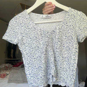 Blommig croppad topp från Hollister - Söt croppad topp från Hollister i vitt med små blå blommor. Toppen har korta ärmar, v-ringning och en dragsko framtill som ger en snygg rynkad effekt. Perfekt för varma dagar och enkel att matcha med jeans eller kjol.