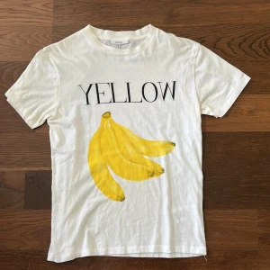Stilren Vit t-shirt med tryck från GANNI - Vit t-shirt från Ganni med tryck av gula bananer och texten 'YELLOW' på framsidan. Klassisk passform och rund halsringning. Tillverkad i 100% bomull för en mjuk och skön känsla.