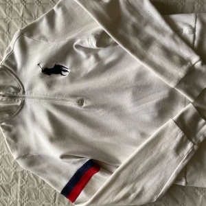 Vit ziptröja från Polo Ralph Lauren - Vit långärmad ziptröja från Polo Ralph Lauren med stor marinblå logga på bröstet. Tröjan har dragkedja framtill och en röd samt marinblå rand på ena ärmen. Perfekt för en sportig och stilren look.