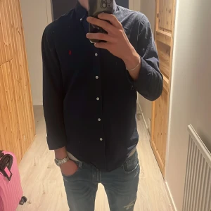 Mörkblå Ralph lauren skjorta  - Snygg mörkblå skjorta med knappar framtill. Perfekt för en stilren look. Skjortan har långa ärmar och en klassisk krage. En defekt som knappt syns när du har den på. Se bild 3 