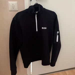 Svart tröja med dragkedja från Boss - Snygg svart tröja från Boss med vit dragkedja och logga på bröstet. Tröjan har en praktisk ficka med dragkedja på armen. Köpt på nk för 2000, men använd inte så många gånger