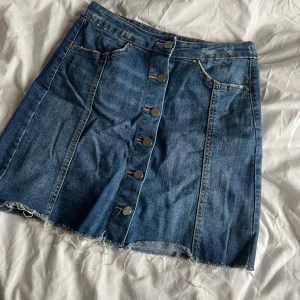 Jeanskjol från Gina tricot - Säljer en klassisk blå jeanskjol med knappar framtill från Gina tricot. Kjolen har rå kant nertill och fickor fram. Perfekt för dig som gillar en tidlös denimstil.