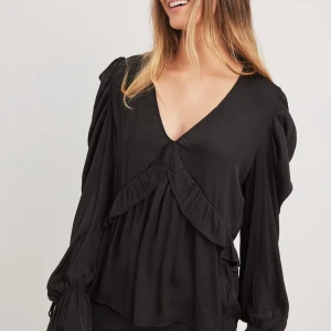 Svart blus med volang och v-ringning - Säljer en svart blus med djup v-ringning och volangdetalj framtill. Blusen har långa ärmar med knyt vid ärmslut. Helt oanvänd och prislappen är kvar. 