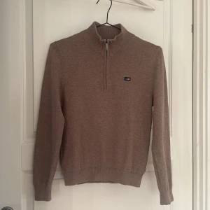 Beige tröja med dragkedja från Lexington - Säljer en stilren beige tröja från Lexington med halv dragkedja och ribbad krage. Perfekt för en avslappnad men ändå snygg look. Tröjan har långa ärmar och en liten logotyp på bröstet.