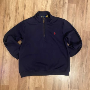 Mörkblå halvzip sweatshirt från Polo Ralph Lauren - Säljer en mörkblå sweatshirt från Polo Ralph Lauren med halv dragkedja och röd broderad logga på bröstet. Tröjan har lång ärm och ribbade muddar vid ärmslut och nederkant. Perfekt för en stilren och avslappnad look.