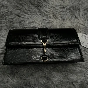 Svart axelväska med ormskinnsmönster - Snygg svart axelväska i syntetmaterial med ormskinnsmönster och guldfärgade metalldetaljer. Väskan har ett lock med spänne och magnetknapp samt justerbar axelrem. Helt oanvänd med prislappen kvar, tyvärr inte med originalförpackningen.