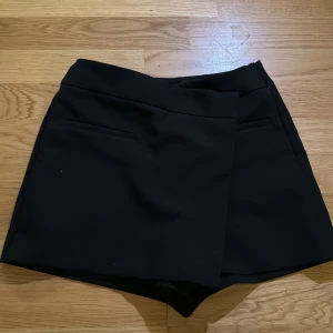 Svart omlottkjol från Zara - Säljer en svart omlottkjol från Zara med stilren design och diskreta fickor framtill. Kjolen har en kort längd och passar perfekt till många olika outfits. Enkel att matcha och superbekväm.