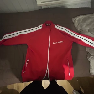 Röd track jacket från Palm Angels - Palm Angels röd track jacket med vita ränder längs ärmarna och vit logga på bröstet. Jackan har dragkedja framtill och två fickor med dragkedja. Sportig och snygg design med hög krage. (Kom med bud)