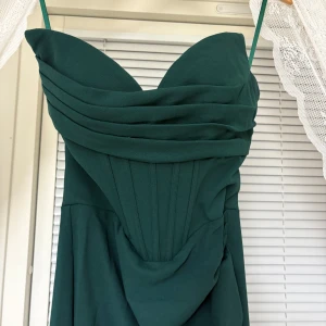 Mörkgrön långklänning  - Elegant mörkgrön långklänning med draperad överdel. Klänningen har snörning i ryggen för en justerbar passform och en figurnära siluett upptill. Perfekt för bal eller festliga tillfällen. Helt ny aldrig använd.