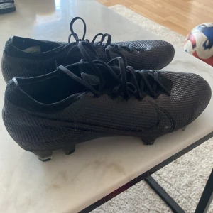 Nike mercurial vapor 13 elite FG  - Säljer ett par svarta Nike fotbollsskor med snörning och rund tå. Skorna har en syntetisk ovandel med diskret struktur och dobbar för spel på gräs. Perfekta för dig som vill ha stilrena och lätta skor på planen.