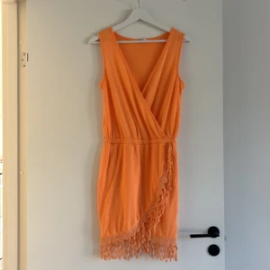 Orange ärmlös klänning med fransar - Snygg orange ärmlös klänning med omlott-design och fransdetaljer längs fållen. Perfekt för sommarens fester. Klänningen har en elastisk midja för en bekväm passform.