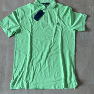 Grön pikétröja från Ralph Lauren - Snygg grön pikétröja från Ralph Lauren med klassisk krage och korta ärmar. Tröjan har en broderad logotyp på bröstet och knappar vid halsen. Perfekt för en stilren look.