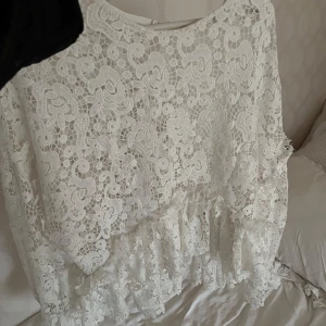 Vit spets blus - Superfin vit blus i spets med volangdetaljer nertill. Blusen har rund halsringning och är täckt av ett vackert blommigt spetsmönster. Perfekt för sommarens tillfällen eller studentfirande.