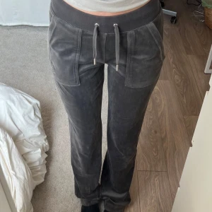 Grå velourbyxor från Juicy Couture - Mysiga grå velourbyxor från Juicy Couture med dragsko i midjan och broderad JC-logga vid fickan. Byxorna har raka ben, fickor fram och bak samt mjukt material som känns lyxigt mot huden.
