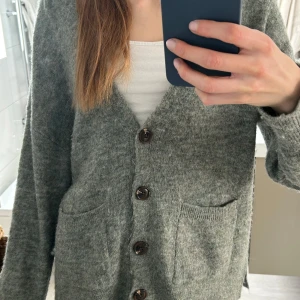 Grå/grön kofta med knappar - Mysig grå/grö kofta med stora bruna knappar och två framfickor. Använd en gång så i perfekt skick! En del alpacka i så ganska varm.