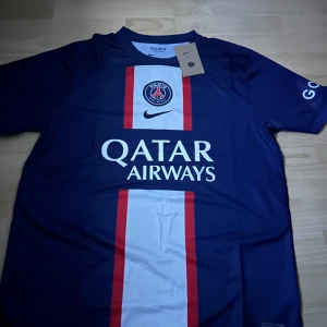 Blå PSG fotbollströja - Säljer en blå helt ny PSG fotbollströja från Nike med korta ärmar. Tröjan har ett vitt och rött vertikalt mönster på framsidan och Qatar Airways som sponsorlogga. Perfekt för fotbollsfans!💯(skriv privat för mer info)
