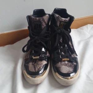 Svarta och ormmönstrade sneakers från Ted Baker - Säljer ett par höga sneakers från Ted Baker med svart lackdetalj, ormmönstrad ovandel och guldfärgade detaljer. Skorna har svarta snören och en vit/rosa sula med Ted Baker-logga undertill. Perfekta för dig som vill sticka ut med din stil.