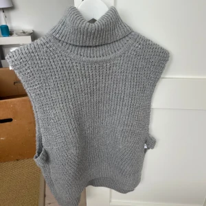 Grå stickad väst med polokrage från Neo Noir - Säljer en grå stickad väst från Neo Noir med hög polokrage. Västen har en grovstickad struktur och är ärmlös, perfekt att bära över en skjorta eller tröja för extra värme och stil.