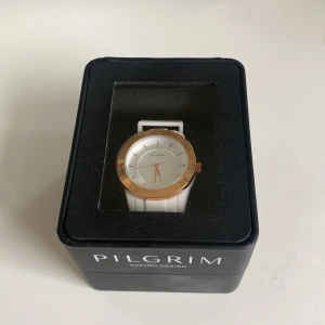 Vit klocka från Pilgrim med gulddetaljer - Snygg klocka från Pilgrim med en rund urtavla. Klockan har guldfärgad kant runt urtavlan och stilrena detaljer. Perfekt accessoar för att lyfta din stil. Med skyddsplasten kvar. Klockan är i sin original förpackning och helt oanvänd.