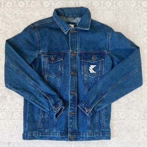 Blå jeansjacka från Karl Kani - Säljer en klassisk blå jeansjacka från Karl Kani i storlek XS. Jackan har knappar framtill, två bröstfickor med lock och broderad logga på ena fickan samt stor KARL KANI-text på ryggen. Perfekt för dig som gillar streetwear.