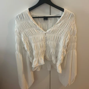 Vit blus med smock från Gina Tricot - Säljer en vit blus från Gina Tricot med smockdetaljer och knappar framtill. Blusen har långärmade ärmar och en v-ringad halsringning. Materialet känns lätt och luftigt, perfekt för vår och sommar. Storlek S