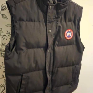 Svart dunväst från Canada Goose - Säljer en svart dunväst från Canada Goose med klassisk logotyp på bröstet. Västen har hög krage, knappar och dragkedja framtill samt två fickor. 