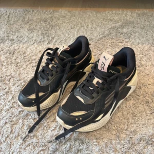 Svarta sneakers från Puma - Snygga svarta sneakers från Puma med vita och beige detaljer. Skorna har en modern design med snörning och orginalsulan är utbytt. Säljer då de inte används mycket