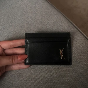 Svart kortfodral från Saint Laurent - Säljer ett stilrent svart kortfodral från Saint Laurent i skinn med guldigt YSL-logo på framsidan. Perfekt för att hålla ordning på dina kort i en smidig och lyxig accessoar. Har lådan 