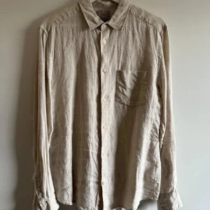 Beige linneskjorta  - Säljer en stilren beige skjorta från H&M i linne. Skjortan har en klassisk krage och knappar framtill. Perfekt för en avslappnad look med en bröstficka som extra detalj. Regular fit för en bekväm passform.