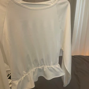 Vit topp med volang - Säljer en vit topp med långa ärmar och volangdetalj nedtill. Toppen har en rund halsringning och är tillverkad i ett mjukt material. Perfekt för dig som gillar stilrena plagg med en liten twist.