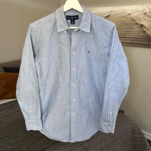 Ljusblå skjorta från Ralph Lauren - Klassisk ljusblå skjorta från Ralph Lauren med button-down krage och broderad logga på bröstet. Skjortan har långa ärmar och knäppning framtill. Perfekt för en stilren och tidlös look.