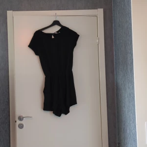 Svart kortärmad jumpsuit - Svart jumpsuit med korta ärmar och rund halsringning. Modellen har en bröstficka och sidofickor. Plagget är tillverkad i ett mjukt material. Perfekt för en avslappnad och stilren look.