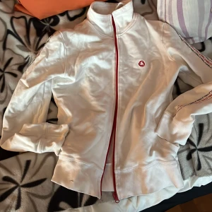 Vit tröja  från Etirel - Säljer en vit jacka från Etirel med röd dragkedja och logga. Jackan har långa ärmar och ribbade muddar. Perfekt för en sportig look.