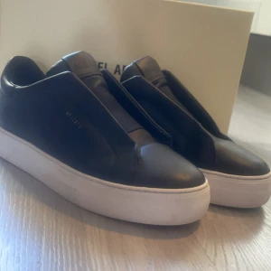 Svarta sneakers från Axel Arigato - Säljer ett par svarta sneakers från Axel Arigato med stilren design och vit sula. Skorna är i skinn och har en minimalistisk look utan synlig snörning, perfekt för dig som gillar cleana och moderna sneakers. Skorna är i fint skick och köptes i februari. Minimalt slitage vilket kan ses på vänster sko. Nypris 2500. Vårt pris 1500kr. Kan skickas med köparen står för porto. 