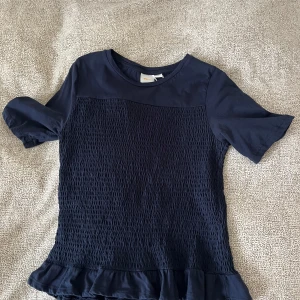 Mörkblå topp med volang från Gina Tricot - Säljer en mörkblå topp från Gina Tricot med korta ärmar och smockad framsida. Toppen har en söt volang längst ner och rund halsringning. Också bara använd 1 gång pga att den är för kort
