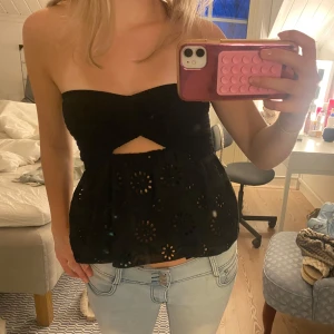 Svart axelbandslös tubtopp med broderade detaljer - Säljer en svart axelbandslös tubtopp med broderade blommönster och liten cut-out framtill. Toppen har en volangliknande nederdel och är perfekt för sommaren. Är i storlek S💗