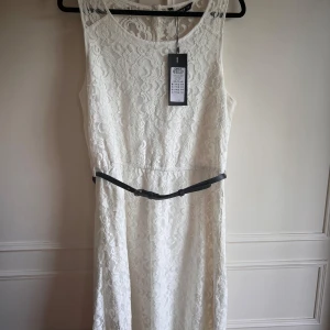 Vit spetsklänning från ONLY, NWT - Elegant vit spetsklänning från ONLY med ärmlös design och ett smalt svart bälte i midjan. Klänningen har en vacker spetsdetalj och knappar i ryggen för en stilren look. Perfekt för sommarens festligheter.