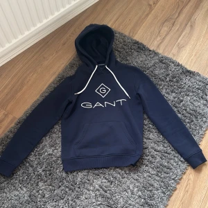 Mörkblå hoodie från GANT - Säljer en mörkblå hoodie från GANT med vit logga och snören. Tröjan har en stor ficka framtill och huva. Perfekt för en avslappnad och stilren look. Köpt på Hede Fashion outlet påse och kvitto finns. Köpt för 1700
