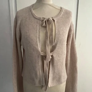 Beige ribbad kofta med rosetter - Säljer en beige ribbad kofta med långa ärmar och två knyt framtill. Tröjan har en mjuk, stickad känsla. Perfekt för lager-på-lager och enkel att styla till olika outfits!💕