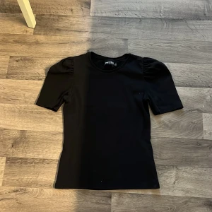 Svart ribbad topp från Pieces - Svart ribbad topp från Pieces med puffiga axlar och korta ärmar. Snygg rund halsringning och stretchigt material som sitter skönt. Perfekt för dig som gillar en enkel men trendig look.